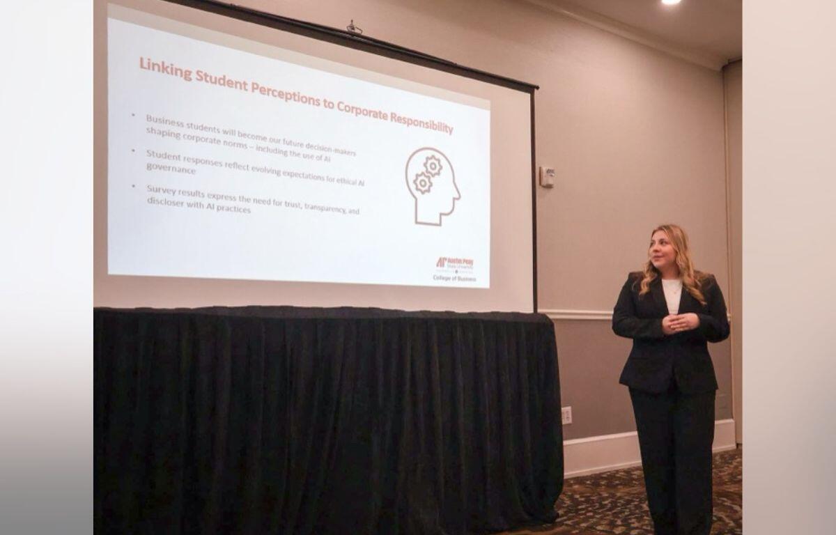Presenter in a black suit standing beside a projected slide titled 'Linking Student Perceptions to Corporate Responsibility' in a conference room.