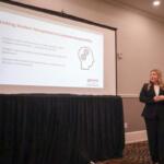 Presenter in a black suit standing beside a projected slide titled 'Linking Student Perceptions to Corporate Responsibility' in a conference room.