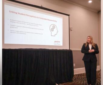 Presenter in a black suit standing beside a projected slide titled 'Linking Student Perceptions to Corporate Responsibility' in a conference room.
