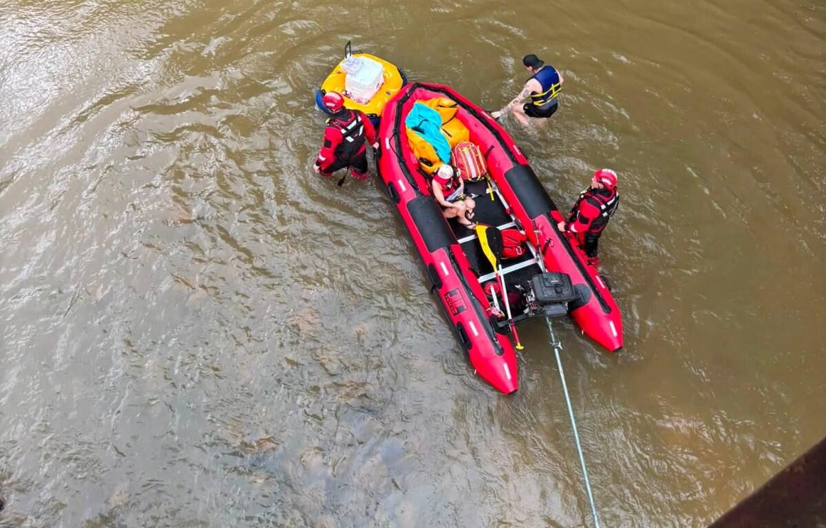 Red inflatable rescue boat with four people in life jackets in a murky river; rescuers assist a swimmer while a rope extends from the boat.