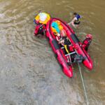 Red inflatable rescue boat with four people in life jackets in a murky river; rescuers assist a swimmer while a rope extends from the boat.