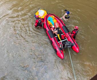 Red inflatable rescue boat with four people in life jackets in a murky river; rescuers assist a swimmer while a rope extends from the boat.