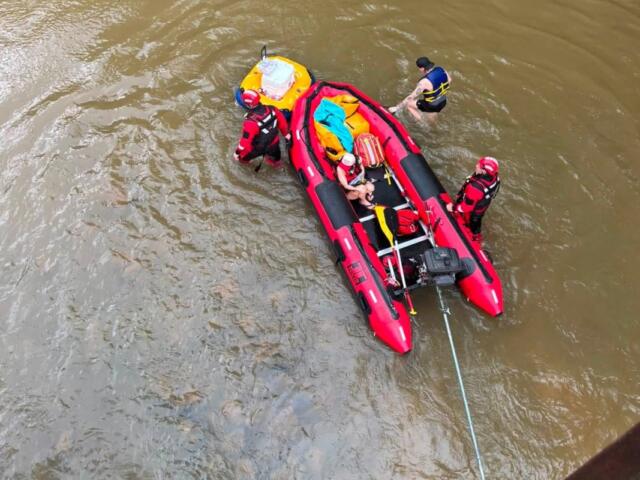 Red inflatable rescue boat with four people in life jackets in a murky river; rescuers assist a swimmer while a rope extends from the boat.