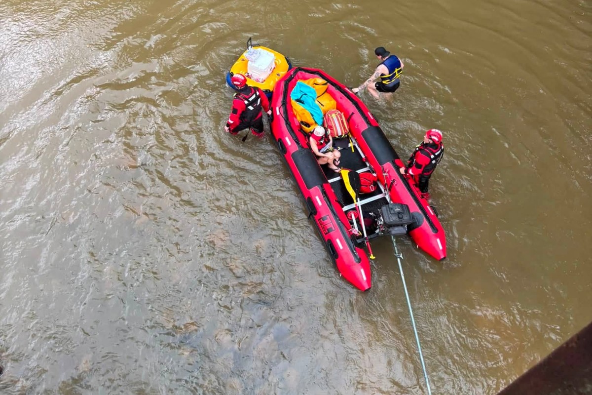 Red inflatable rescue boat with four people in life jackets in a murky river; rescuers assist a swimmer while a rope extends from the boat.