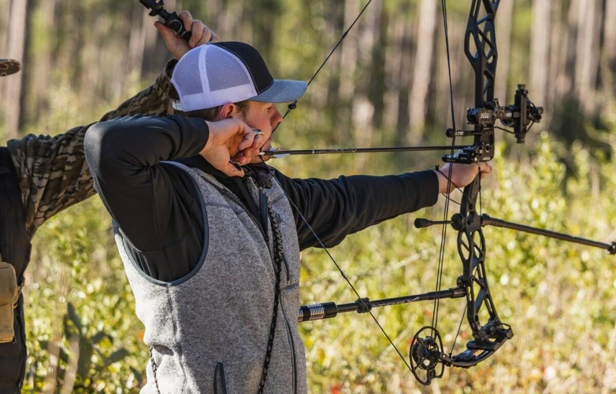 Archer with a drawn bow aiming in a sunlit forest, wearing a gray vest and cap.