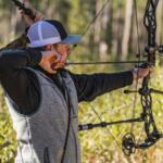 Archer with a drawn bow aiming in a sunlit forest, wearing a gray vest and cap.
