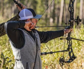 Archer with a drawn bow aiming in a sunlit forest, wearing a gray vest and cap.