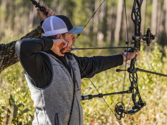 Archer with a drawn bow aiming in a sunlit forest, wearing a gray vest and cap.