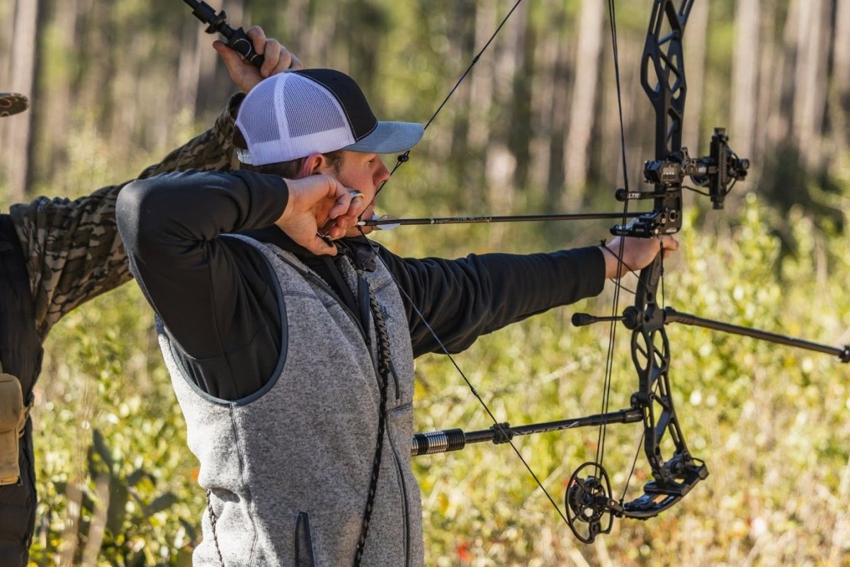 Archer with a drawn bow aiming in a sunlit forest, wearing a gray vest and cap.
