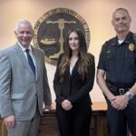 Three people pose in front of the Clarksville, TN city seal: a man in a gray suit, a woman in a black blazer, and a uniformed officer.