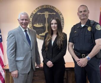 Three people pose in front of the Clarksville, TN city seal: a man in a gray suit, a woman in a black blazer, and a uniformed officer.