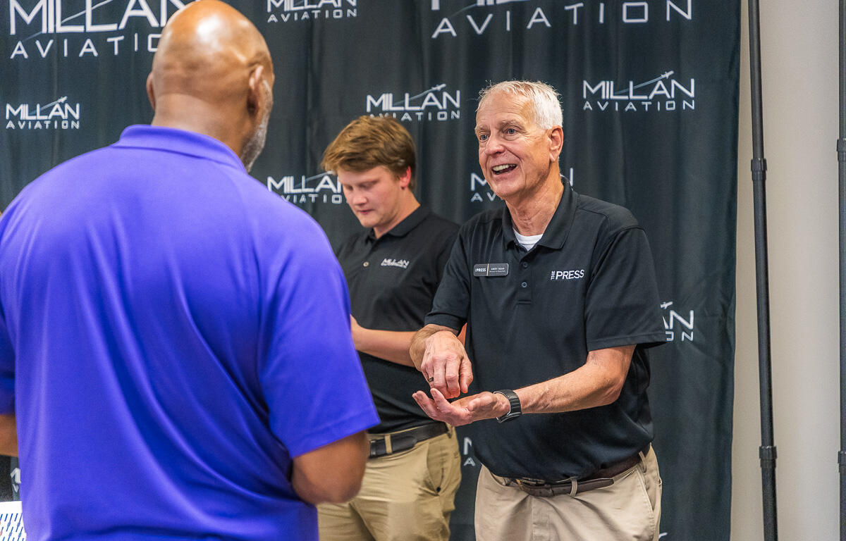Older man in a black polo with a PRESS badge demonstrates a gesture to a man in a blue polo at a Millan Aviation event backdrop.