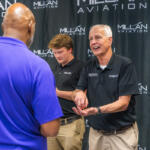 Older man in a black polo with a PRESS badge demonstrates a gesture to a man in a blue polo at a Millan Aviation event backdrop.