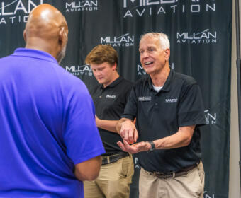 Older man in a black polo with a PRESS badge demonstrates a gesture to a man in a blue polo at a Millan Aviation event backdrop.