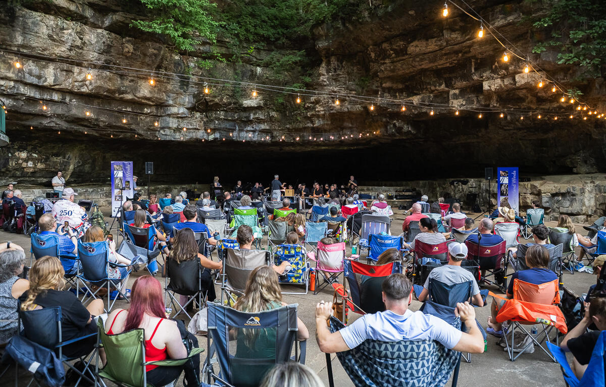 Audience seated in lawn chairs inside a rocky cave, a live band performing on a small stage under string lights.
