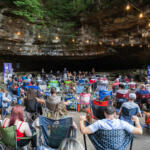 Audience seated in lawn chairs inside a rocky cave, a live band performing on a small stage under string lights.