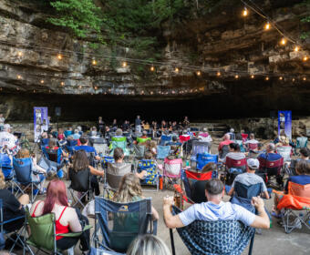Audience seated in lawn chairs inside a rocky cave, a live band performing on a small stage under string lights.