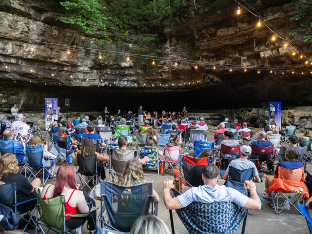 Audience seated in lawn chairs inside a rocky cave, a live band performing on a small stage under string lights.