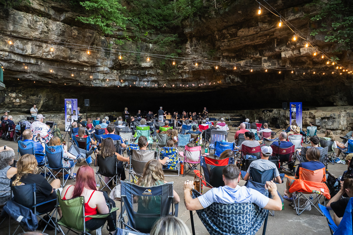 Audience seated in lawn chairs inside a rocky cave, a live band performing on a small stage under string lights.
