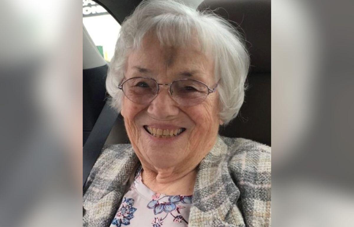 Smiling elderly woman with short white hair and glasses, wearing a plaid jacket in a car seatbelted in the car.