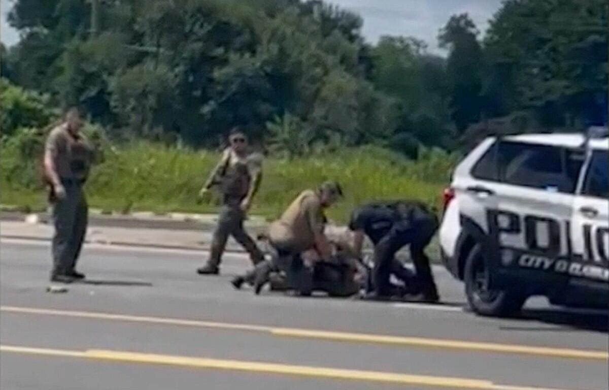 Several officers restrain a person on the road beside a white police SUV, with a bystander watching.