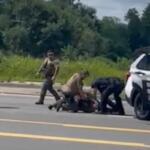 Several officers restrain a person on the road beside a white police SUV, with a bystander watching.