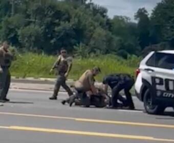 Several officers restrain a person on the road beside a white police SUV, with a bystander watching.