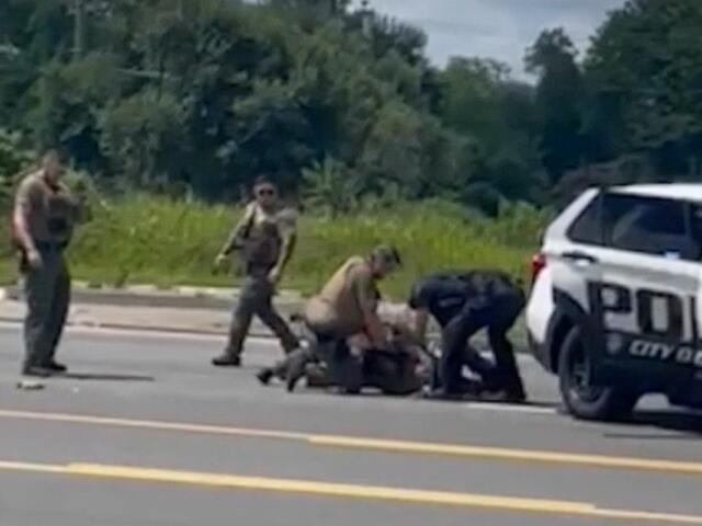 Several officers restrain a person on the road beside a white police SUV, with a bystander watching.