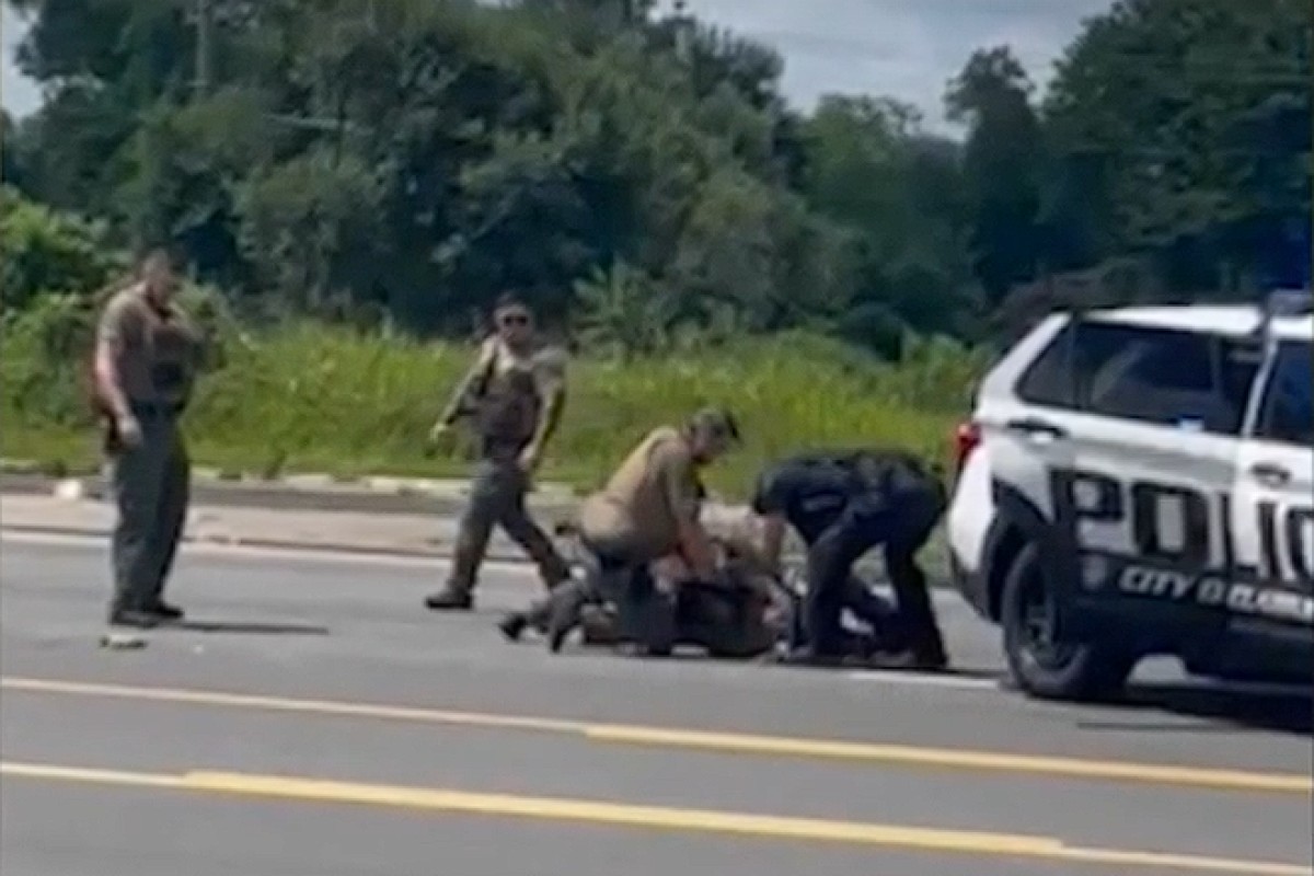 Several officers restrain a person on the road beside a white police SUV, with a bystander watching.