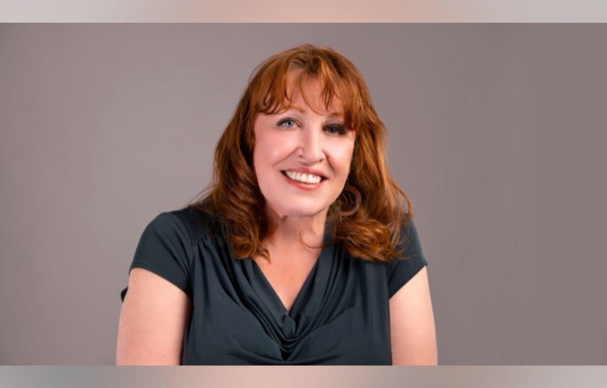 Smiling woman with shoulder-length red hair wearing a dark teal blouse against a neutral gray background the portrait.