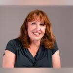 Smiling woman with shoulder-length red hair wearing a dark teal blouse against a neutral gray background the portrait.