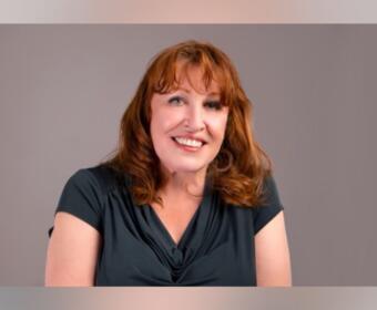 Smiling woman with shoulder-length red hair wearing a dark teal blouse against a neutral gray background the portrait.