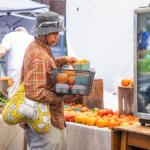 Shopper in a plaid shirt and bucket hat holds a basket of oranges at an outdoor market, with tomatoes and peppers nearby.