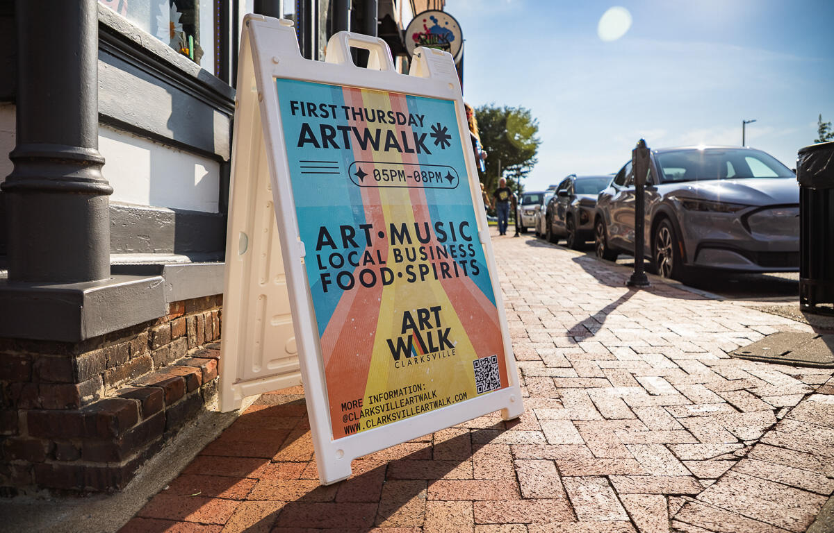 A-frame sign on a sunlit brick sidewalk advertising First Thursday ArtWalk, 05PM–08PM, featuring art, music, local business, and food/spirits.