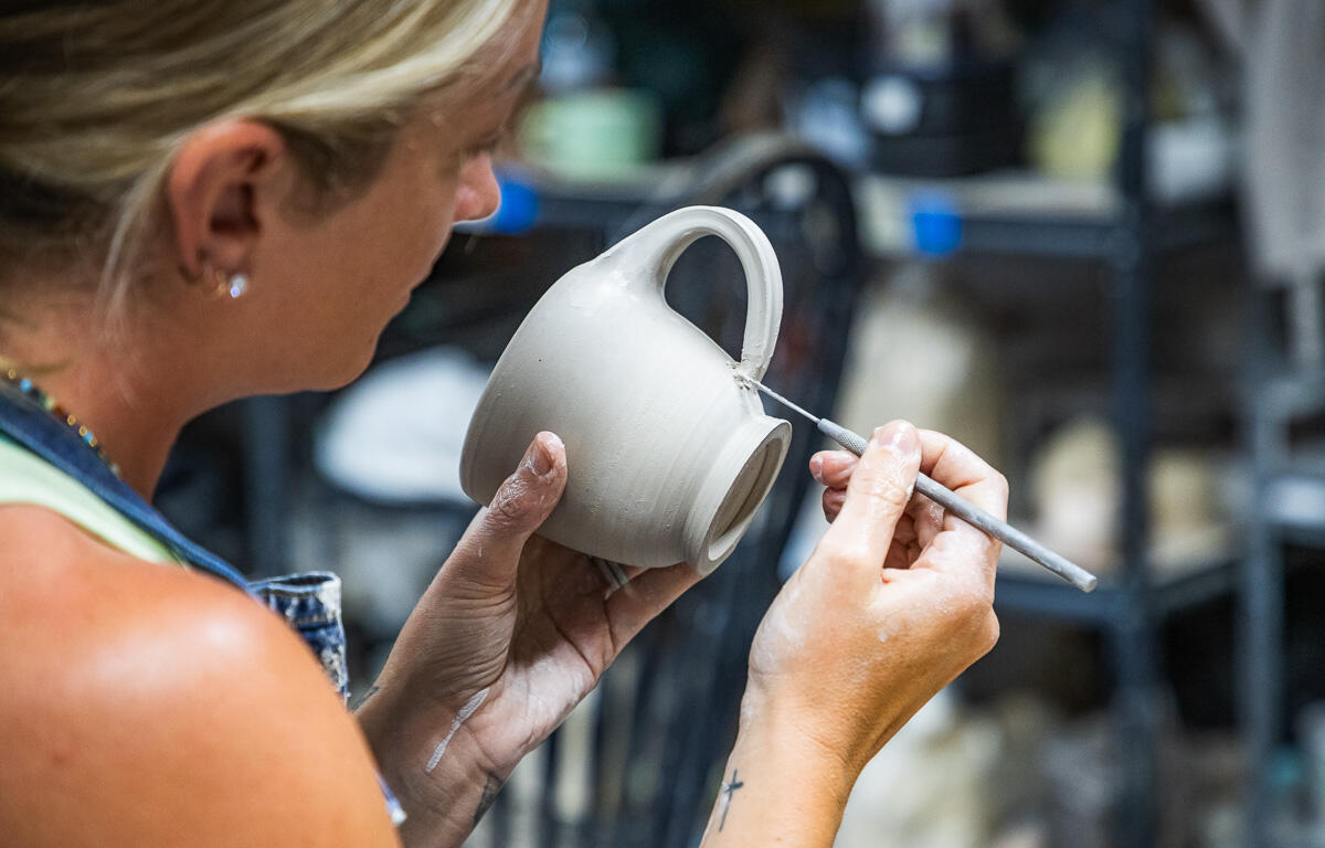 Potter shaping a gray ceramic mug with a sculpting tool in a pottery studio