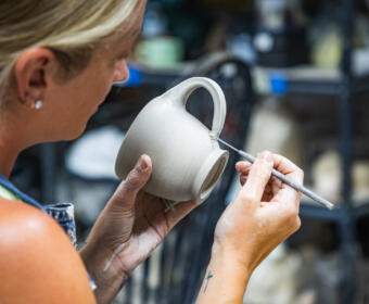 Potter shaping a gray ceramic mug with a sculpting tool in a pottery studio