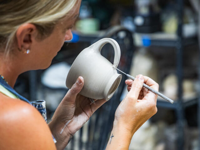 Potter shaping a gray ceramic mug with a sculpting tool in a pottery studio