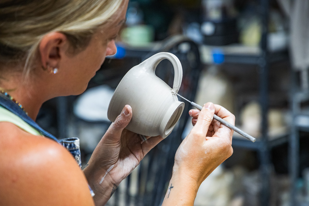 Potter shaping a gray ceramic mug with a sculpting tool in a pottery studio