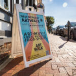 A-frame sign on a sunlit brick sidewalk advertising First Thursday ArtWalk, 05PM–08PM, featuring art, music, local business, and food/spirits.