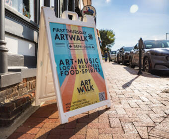 A-frame sign on a sunlit brick sidewalk advertising First Thursday ArtWalk, 05PM–08PM, featuring art, music, local business, and food/spirits.