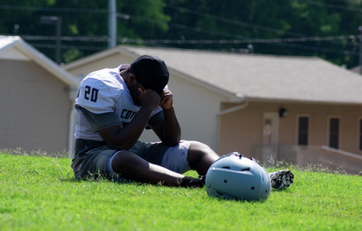 Male American football player in jersey number 20 sits on a grassy field, head in hands, with his helmet on the ground nearby.