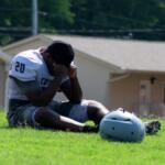 Male American football player in jersey number 20 sits on a grassy field, head in hands, with his helmet on the ground nearby.