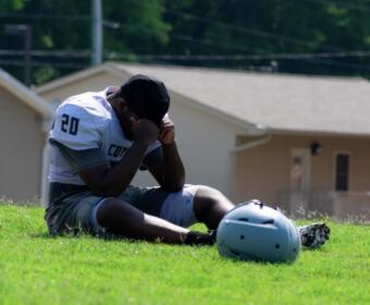 Male American football player in jersey number 20 sits on a grassy field, head in hands, with his helmet on the ground nearby.