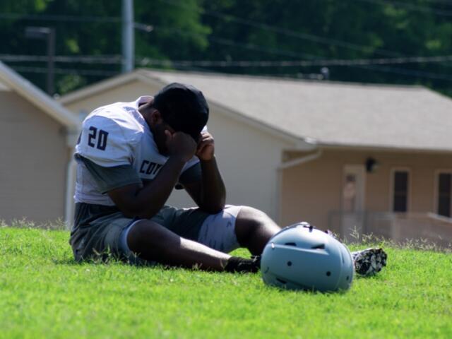 Male American football player in jersey number 20 sits on a grassy field, head in hands, with his helmet on the ground nearby.