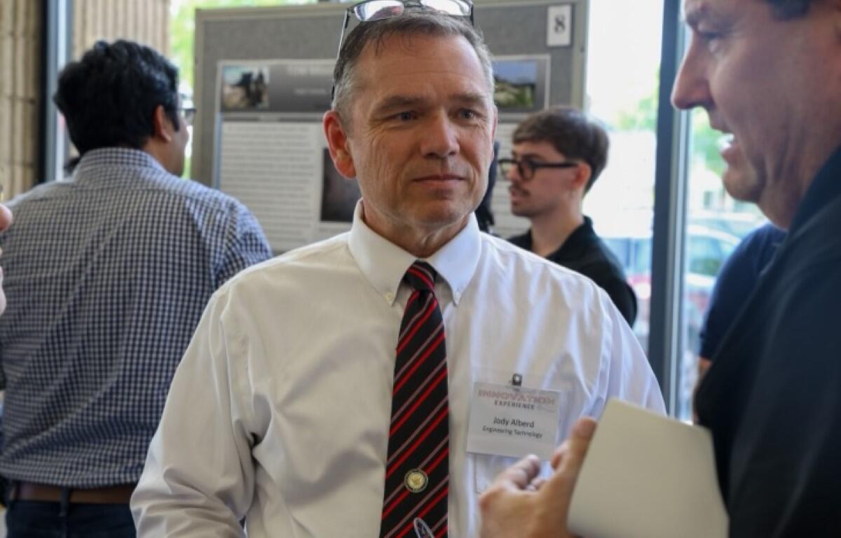 Middle-aged man in a white shirt and red striped tie speaks with a colleague at a conference; name badge visible.