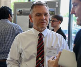 Middle-aged man in a white shirt and red striped tie speaks with a colleague at a conference; name badge visible.