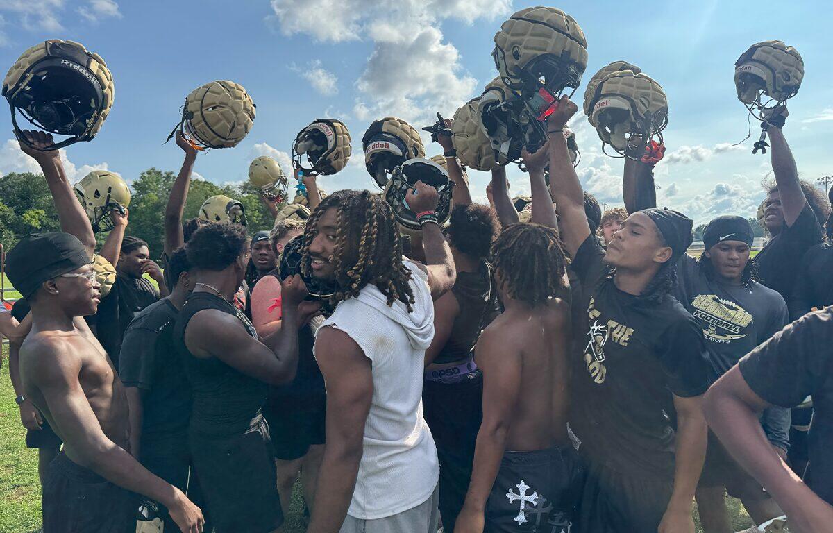 Football players in black uniforms form a huddle, raising gold helmets in celebration on a sunny practice field.