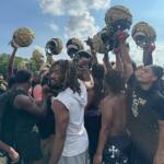 Football players in black uniforms form a huddle, raising gold helmets in celebration on a sunny practice field.