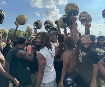 Football players in black uniforms form a huddle, raising gold helmets in celebration on a sunny practice field.