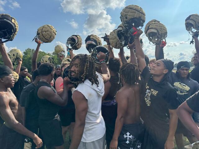 Football players in black uniforms form a huddle, raising gold helmets in celebration on a sunny practice field.